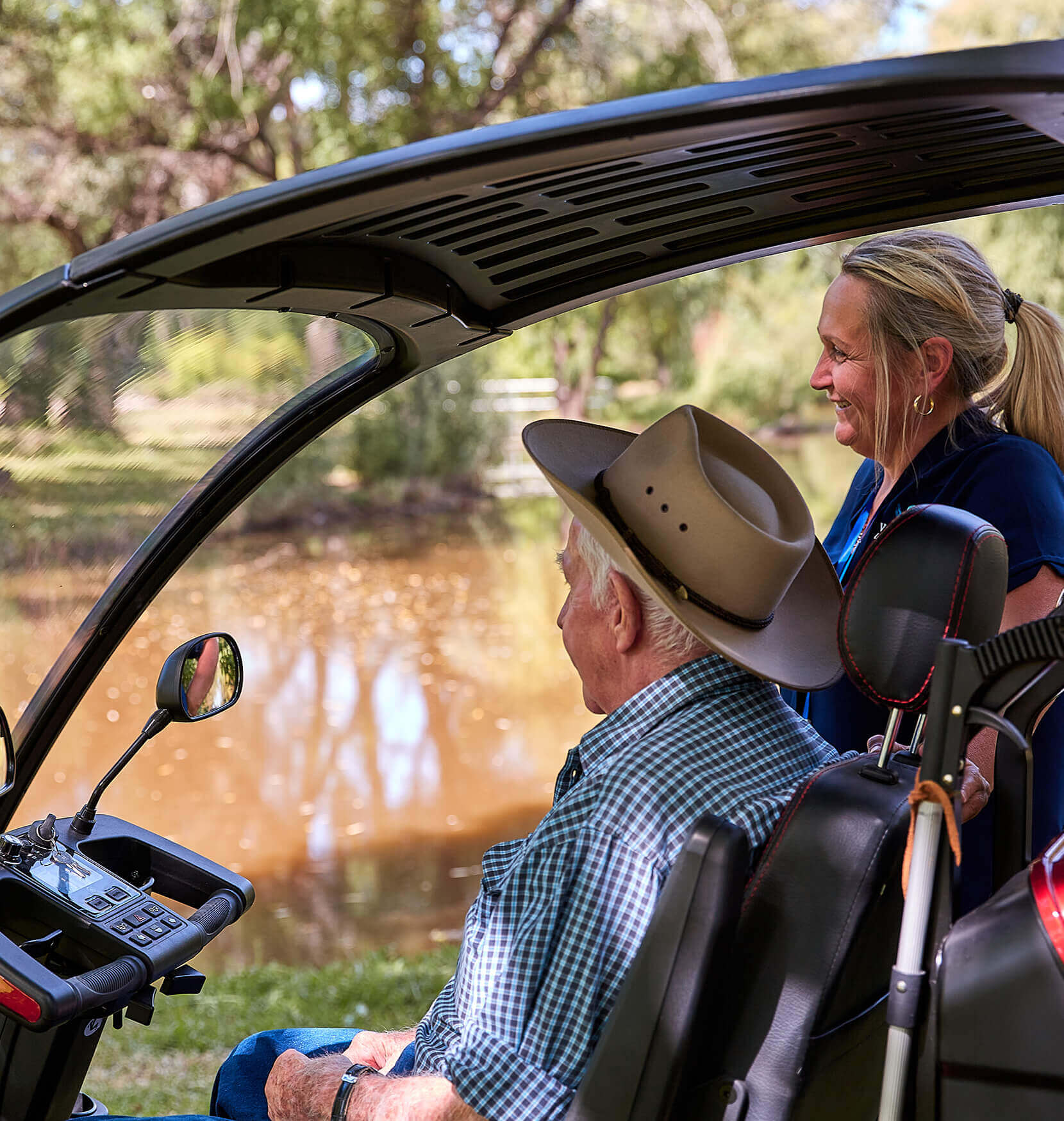 A man in a akubra hat and chequered shirt sitting in the cabin of a buggy gazing out at a muddy river. A woman is standing next to him outside the buggy smiling. She had a blonde hair in a ponytail.